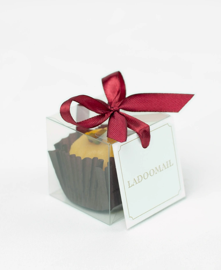 Ladoomail celebration favour: a kesar peda finished with pistachio in a clear favour box tied with a signature burgundy ribbon bow and swing tag.