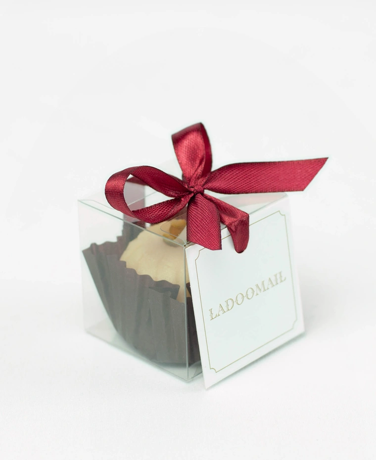 Ladoomail celebration favour: a khoya peda finished with pistachio in a clear favour box tied with a signature burgundy ribbon bow and swing tag.