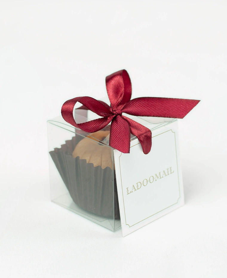 Ladoomail celebration favour: a Mathura peda in a clear favour box tied with a signature burgundy ribbon bow and swing tag.