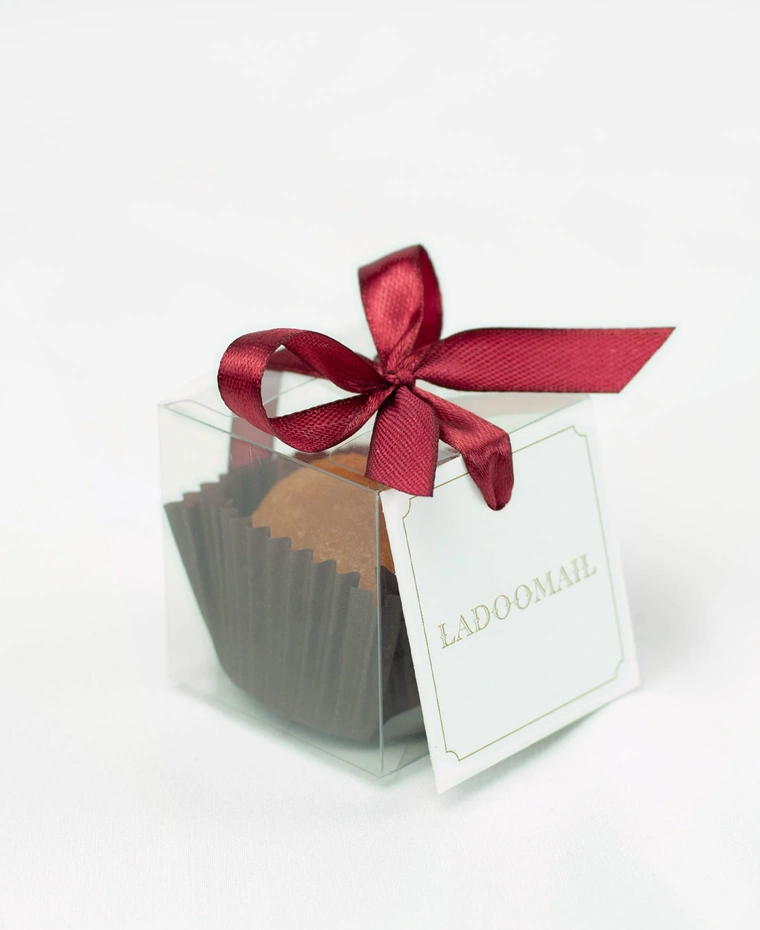 Ladoomail celebration favour: a classic motichoor ladoo in a clear favour box tied with a signature burgundy ribbon bow and swing tag.