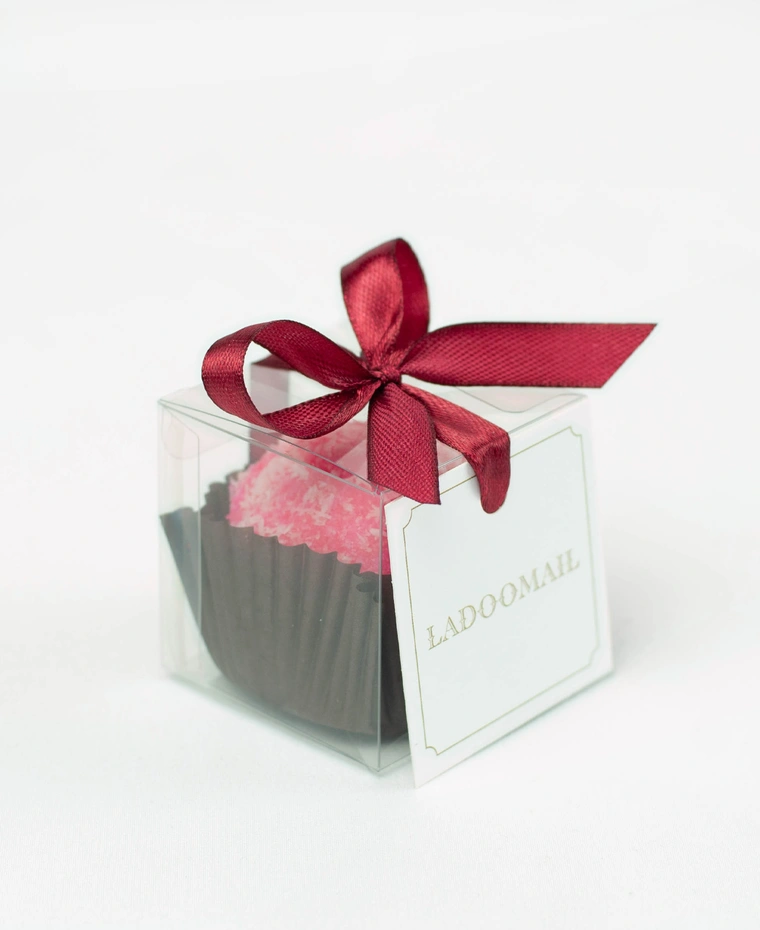 Ladoomail celebration favour: a rose coconut artisan ladoo in a clear favour box tied with a signature burgundy ribbon bow and swing tag.