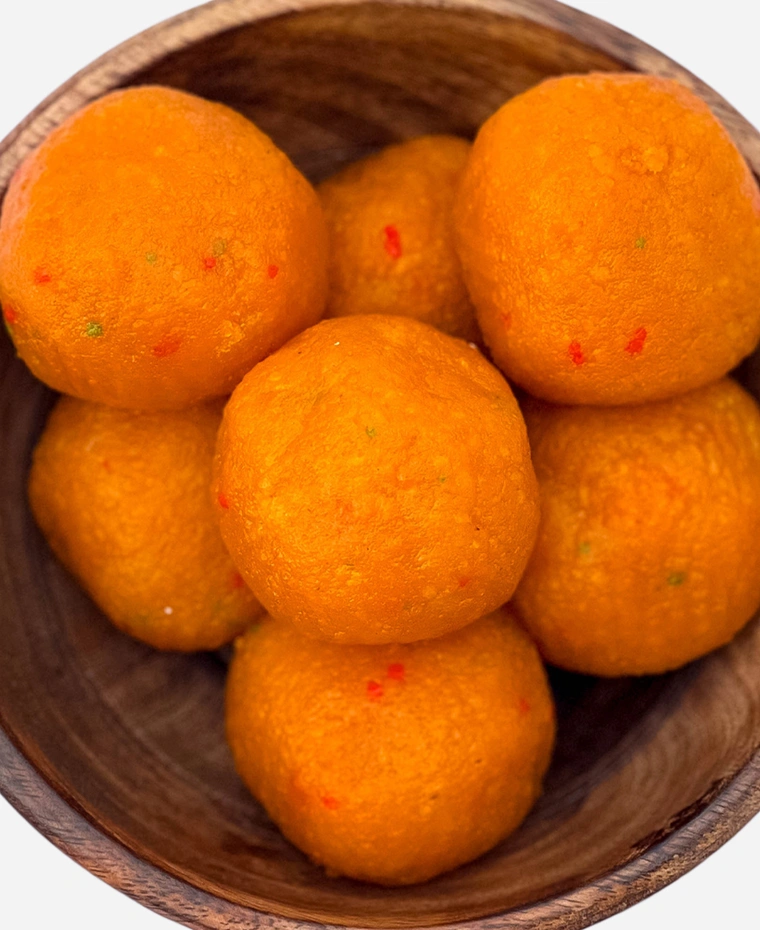 Close-up of handmade orange motichoor ladoo, fine-grained and flecked with red and green, in a wooden bowl.