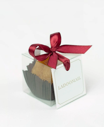 Ladoomail celebration favour: a classic boondi ladoo in a clear favour box tied with a signature burgundy ribbon bow and swing tag.