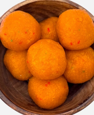 Close-up of handmade orange motichoor ladoo, fine-grained and flecked with red and green, in a wooden bowl.