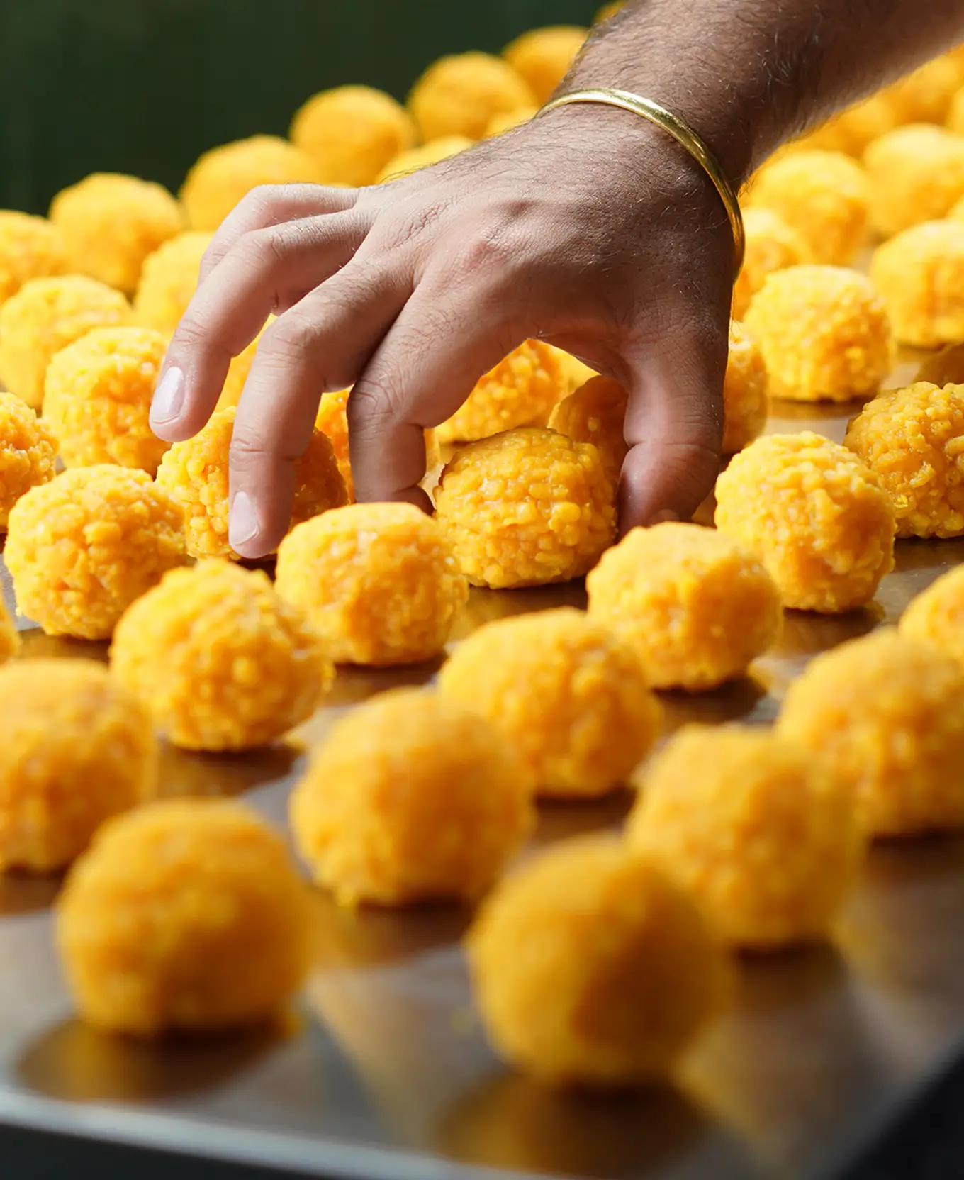 Hand reaching for freshly rolled golden boondi ladoo on a tray.