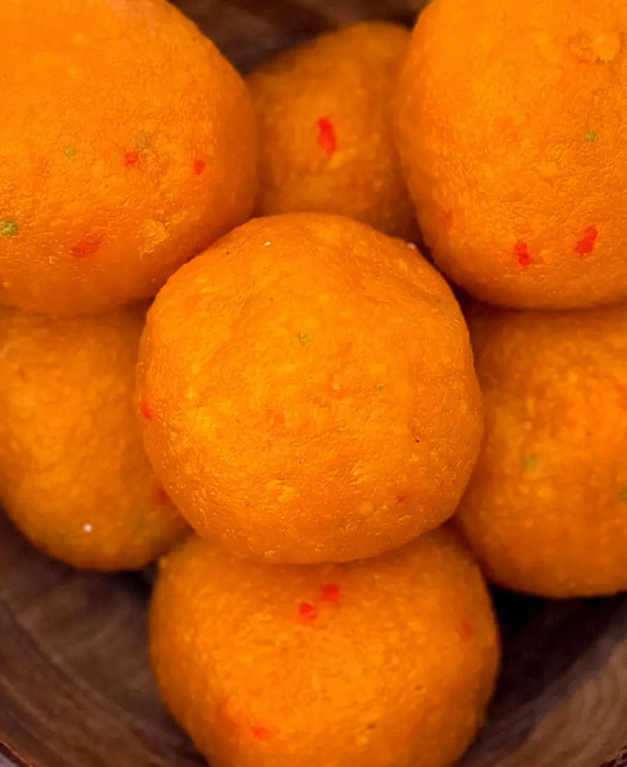 Close-up of handmade orange motichoor ladoo, fine-grained and flecked with red and green, in a wooden bowl.