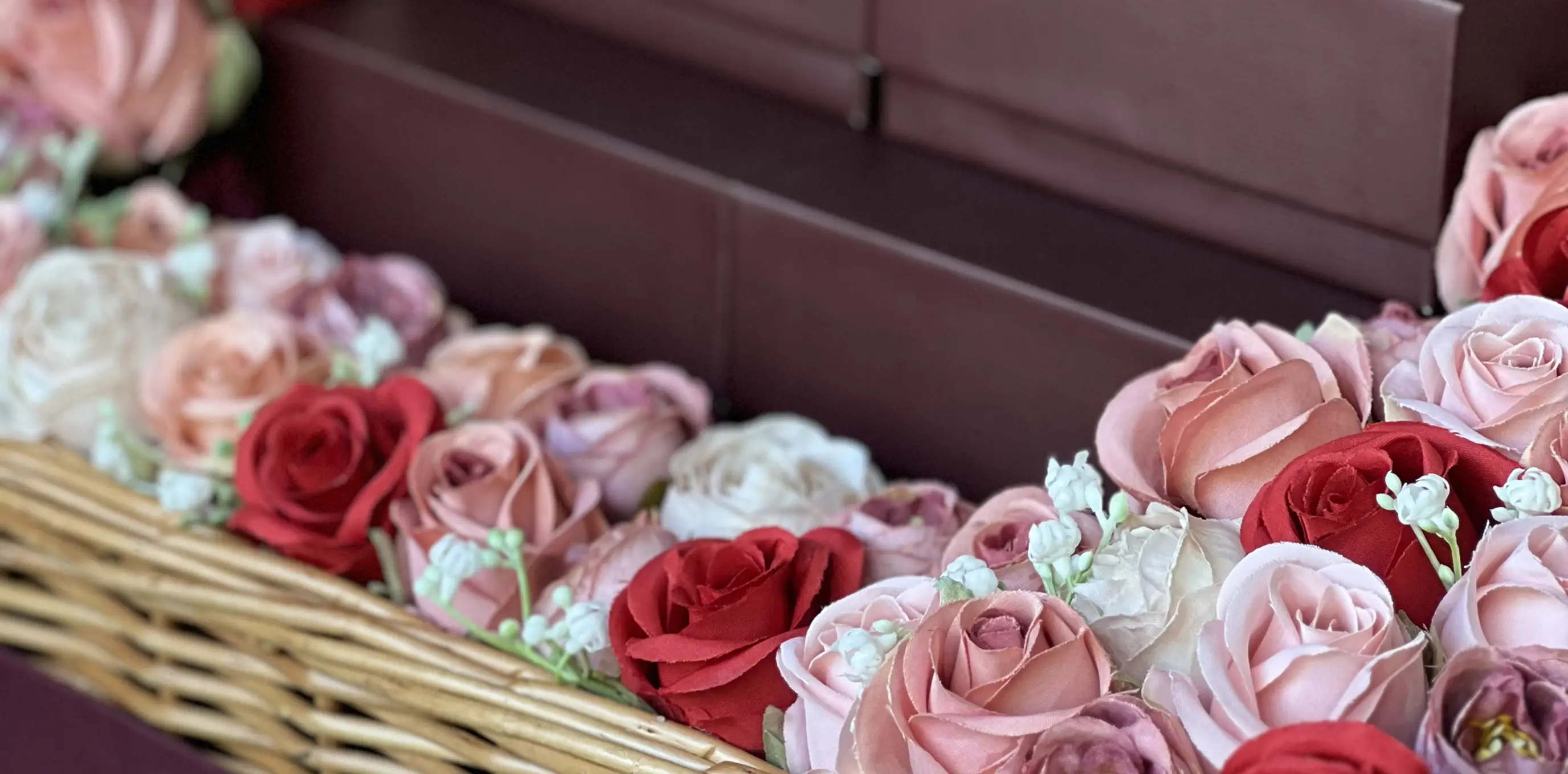 Ladoomail wicker basket of red, pink and ivory silk roses beside stacked burgundy gift boxes.