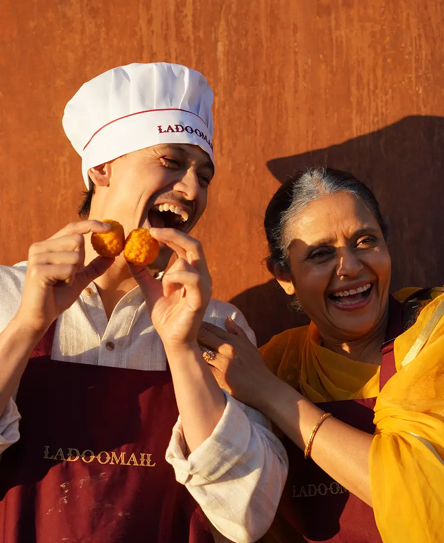 Two people in Ladoomail-branded attire laughing together while sharing a ladoo.