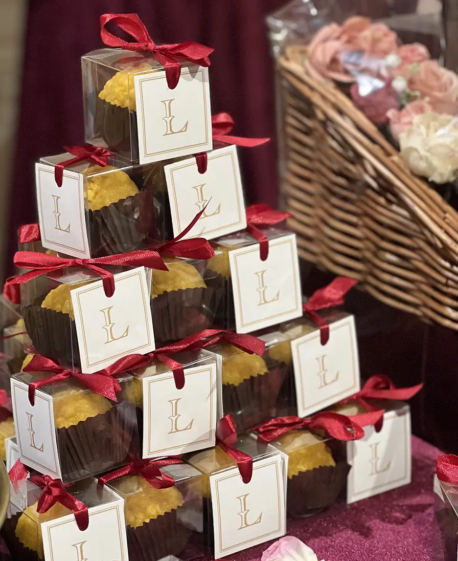 Stacked Ladoomail celebration favours with monogrammed tags and red ribbon beside a flower-filled wicker basket.