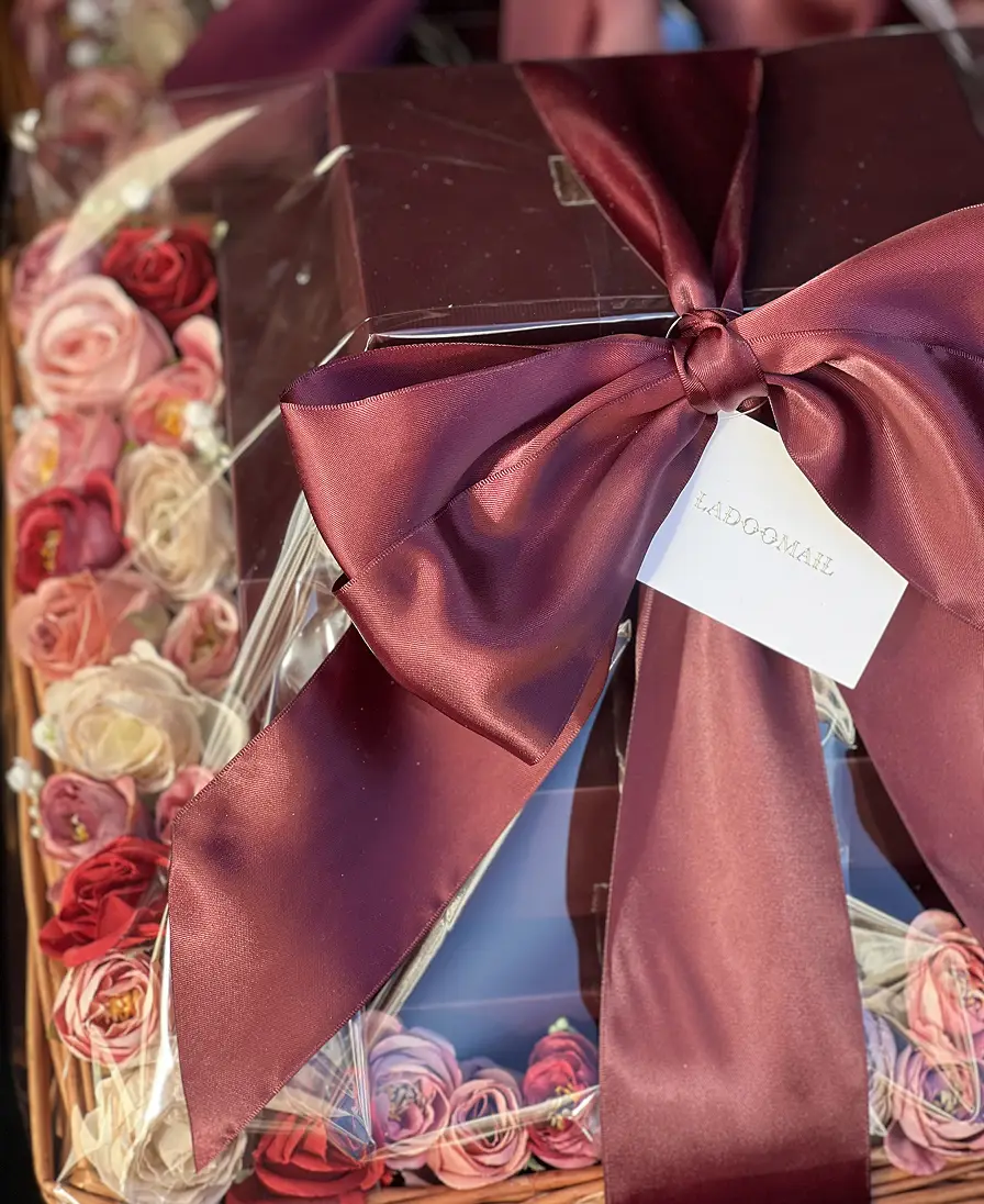 The Gift Basket Ladoomail wicker gift basket lined with flowers, tied with a burgundy bow and branded gift tag.