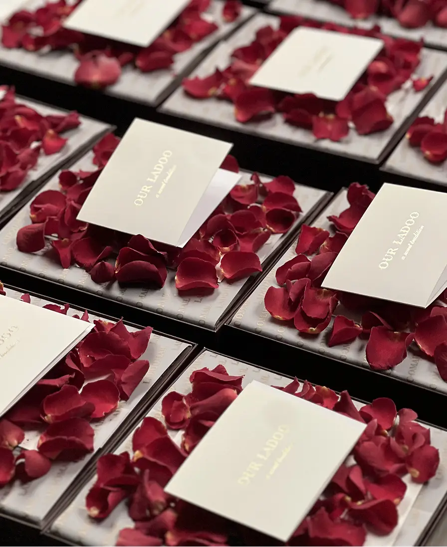 Rows of Ladoomail burgundy gift boxes with 'Our Ladoo' cards and rose petals, prepared as wedding announcements.